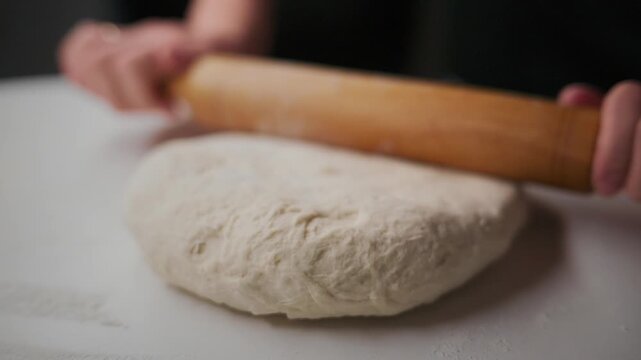 Hands roll dough with wooden roller on white surface. Action shows dough flattening slowly. Focus on rolling motion and texture of dough. Ideal for cooking, baking, or food preparation content