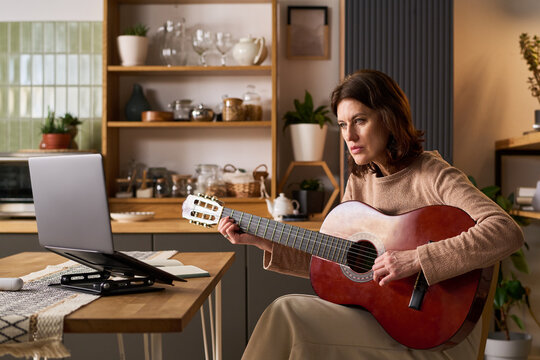 Caucasian middle aged woman playing guitar while following online lesson on laptop at home, learning new skill. Useful for e learning, hobby training, remote education marketing