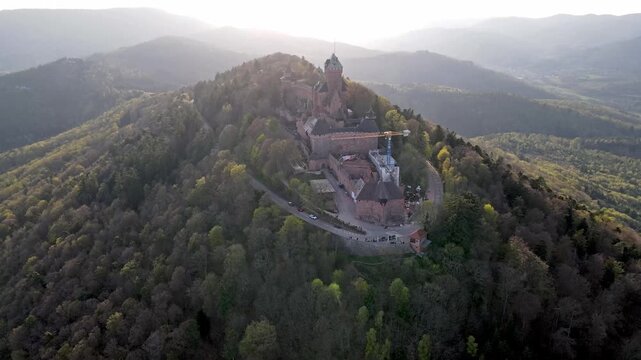 Aerial view of medieval castle on green mountain slope catching peaceful sunrise with morning light filtering through Vosges forest. Winding pathway leading to hilltop fortress with serene dawn