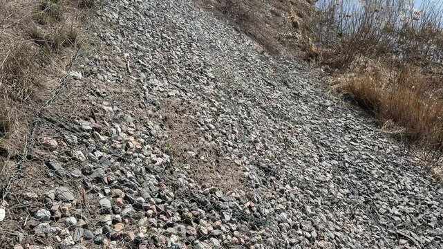 Crushed stone riverbank with dry grass, stainless wire mesh over pebble embankment beside calm river, textured closeup of rocks