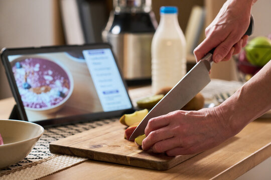 Close up of middle aged woman hands cutting fruit on board while following tablet recipe at home, learning cooking skills online. Ideal for e learning, digital lifestyle, home cooking