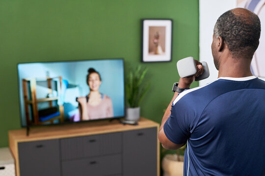 African American senior man exercising with dumbbells while following online fitness class on television at home, supporting active aging, wellness marketing, rear view