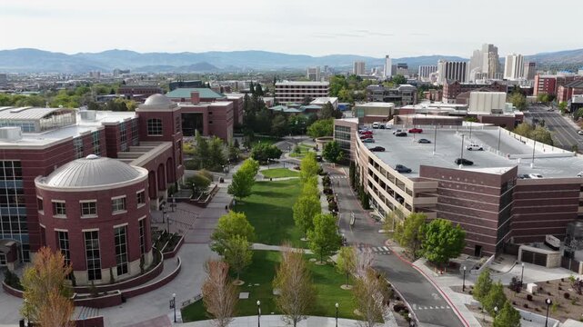 University of Nevada Reno red brick rotunda and tree-lined quad with parking structures, downtown Reno skyline and arid mountains beyond under cloudy and sunny sky. Nevada, USA. Aerial ascend shot.