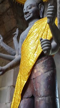 A multi-armed stone statue of a deity, draped in a golden cloth and adorned with a yellow umbrella, stands within the ancient stone ruins of Angkor Wat, Siem Reap, Cambodia.