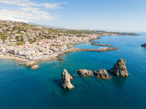 Aerial view of the Islands of the Cyclops sea stacks rising from the turquoise Mediterranean Sea near the coastal town and harbor in Aci Castello, Italy.