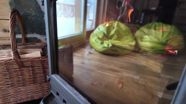 Close‑up of an anthracite cast iron stove with visible flames and cabin reflections, next to a wicker basket of kindling.