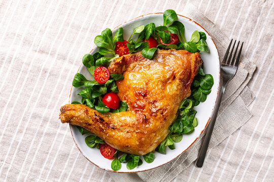 Top view of Dinner or lunch with oven roasted chicken leg quarter and fresh valerian or corn salad with cherry tomatoes on white plate, abobe light linen tablecloth.