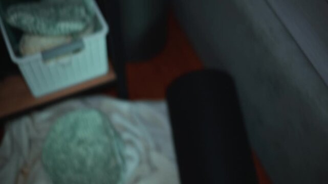 An orange cat approaches a cluttered table in a dimly lit room. The cat jumps down from the table to a nearby chair, navigating through the items scattered around