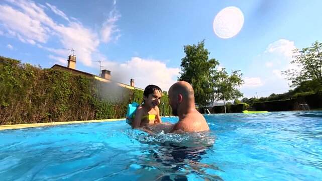 Playful son with arm floaties bravely jumping into the clear blue water of a swimming pool, being caught by his smiling father during a beautiful sunny summer day creating a splash