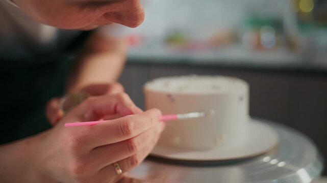 Close-up of hands delicately painting cake with fine brush. Person focuses intensely, ensuring precision on cake surface. Cake rotates slowly on silver stand for even decoration