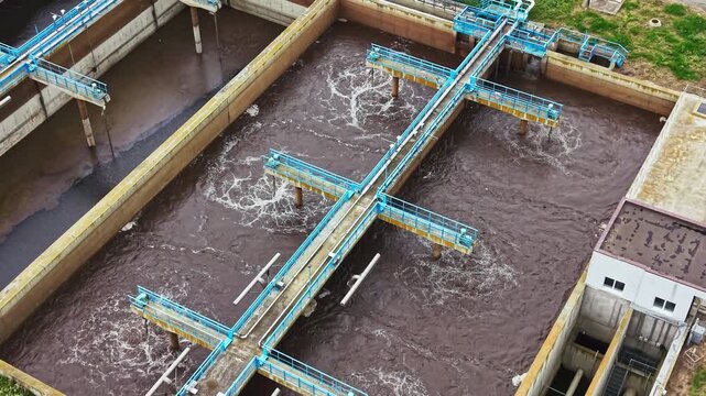 Aerial sewage treatment plant aeration basins bubbling over dark water, drone perspective showing turquoise aerators churning sludge, foam and oxygen rise,
