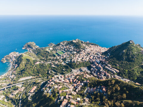 Aerial view of the hilltop village of Castelmola with its historic stone buildings, lush green slopes, and the deep blue Ionian Sea in the background Castelmola, Sicily, Italy.