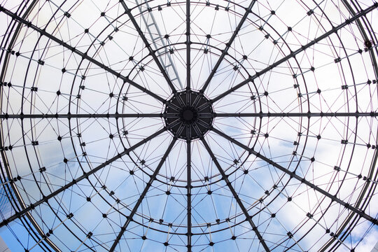 Glass dome architecture spreads a radial geometric pattern overhead, where an urban building roof opens toward sky through a circular grid.