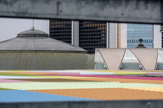 Layered architecture places a colorful roof beneath an urban skyline, where building facade shapes stack across the frame with graphic depth.