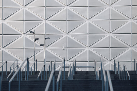 Abstract architecture shows a geometric stairway beneath a patterned facade, where railings and shadow guide an urban building entrance into depth.