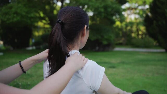 This outdoor scene shows a therapist performing shoulder massage on a female adult with ponytail in a lush park, providing physical relaxation and muscle tension relief.