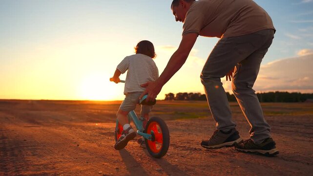 A man assists a young boy riding a bicycle at sunset. The sunset creates a vibrant backdrop as the boy navigates the dirt path on his bicycle.