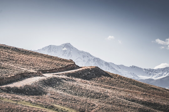 A dusty road on the Pamir Highway winds through mountain hills and slopes among rocks against the backdrop of the Tien Shan mountain peaks