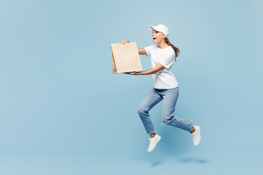 Full body delivery employee woman wear white cap T-shirt uniform work as dealer hold brown blank craft paper takeaway bag mock up jump isolated on plain pastel blue background studio. Service concept.