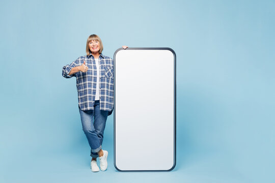 Full body smiling elderly blonde woman 50s years old wear checkered shirt casual clothes big huge blank screen mobile cell phone smartphone with area show thumb up isolated on plain blue background.