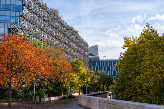 Autumn trees line a Paris France urban park, where a walkway leads past textured architecture and building facades in a layered landscape.