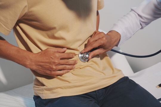 A close-up shot of a doctor examining a patient's stomach area with a stethoscope to listen for bowel sounds or abdominal issues, while the patient rests his hand on his stomach.