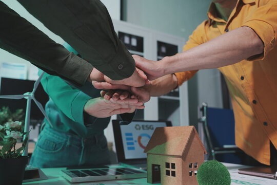 A group of professionals stack their hands together in a sign of unity over a conference table. The scene emphasizes teamwork in green business, featuring a model house, a miniature wind turbine.