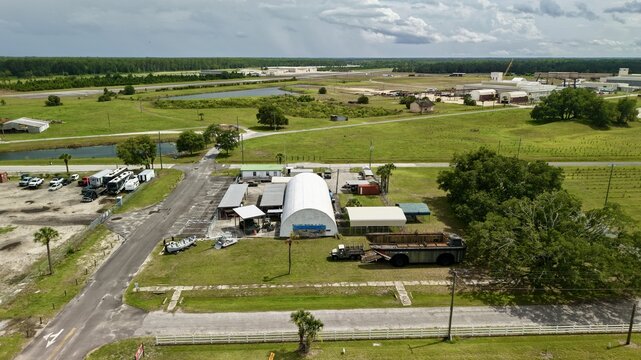 Aerial view of an arched white hangar and a large amphibious vehicle surrounded by green fields and industrial structures in Green Cove Springs, Florida, United States.