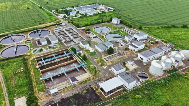 Aerial wastewater treatment plant industrial facility surrounded by green fields, multiple circular clarifiers, rectangular aeration basins, white storage