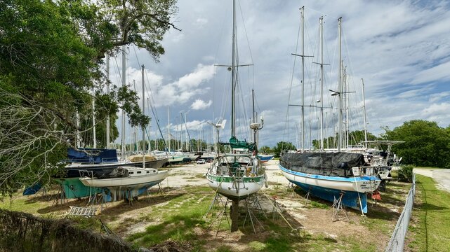 Aerial view of sailboats on stands in a dry dock boatyard surrounded by lush green trees under a cloudy sky in Green Cove Springs, Florida, United States.