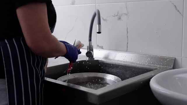 Female kitchen worker wearing blue gloves cuts vegetables with scissors over a stainless steel sink, water splashing, and a white bowl visible in the background