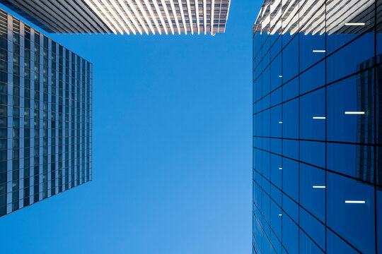 Blue sky cuts between urban skyscraper facades, where glass architecture and geometric lines frame a clean upward view of towers overhead.