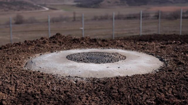 Circular concrete manhole cover embedded in freshly disturbed soil, surrounded by a rural landscape with distant hills and a fence in the background