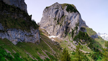 Large rocky mountain stands tall among green hills in the Swiss Alps during daylight hours with some snow patches visible on the slopes © Joachim