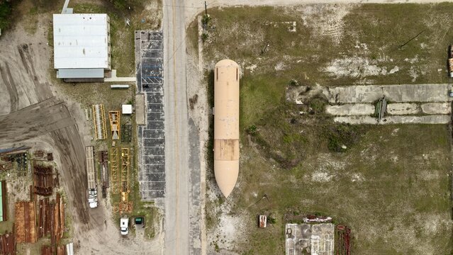 Aerial view of a large orange cylindrical tank or rocket component lying in an industrial storage yard next to a road and parking area in Green Cove Springs, Florida, United States.