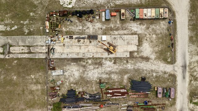 Aerial view of an industrial storage yard featuring a yellow crane, shipping containers, and stacks of pipes on a sandy lot in Green Cove Springs, Florida, United States.