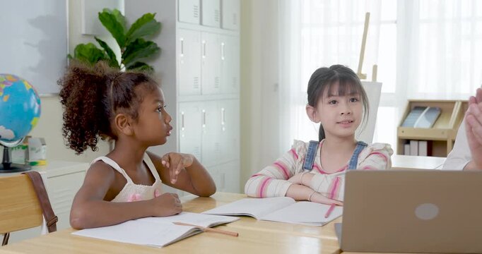 Two elementary girls sit at wooden desk with notebooks and laptop, one girl turns toward classmate during back to school study time, building friendship, listening skill, learning habit, respect