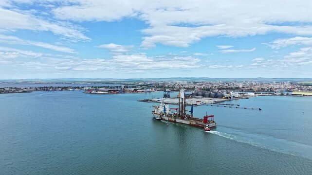 An industrial dredging vessel moves through harbor waters. The city skyline is visible in the background with various structures along the shore on a clear day.