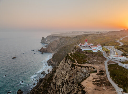 Aerial view of Cape Roca Lighthouse perched on rugged cliffs overlooking the Atlantic Ocean during a golden sunset in Colares, Lisboa, Portugal.