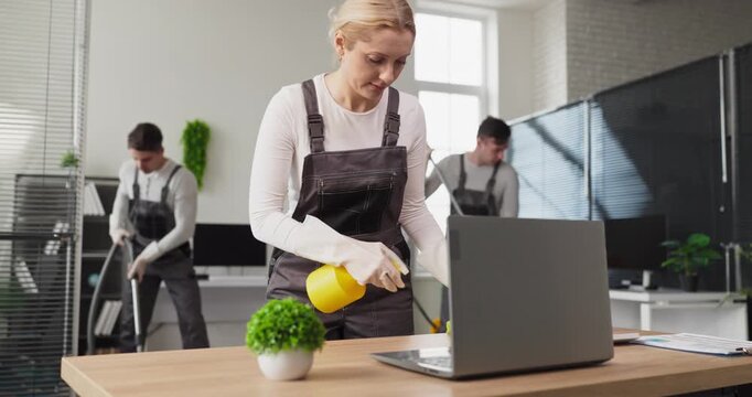 Cleaner team dusting office surfaces for sanitation. Janitor team uniformed wiping desk and laptop. Professional housekeeping service uses spray and cloth. Clean office hygiene and safety concept.