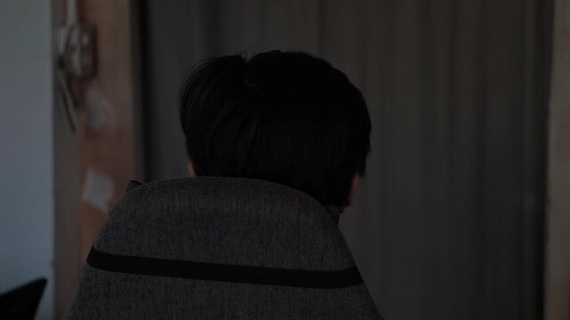 Rear static shot of a man sitting down on an ergonomic chair and resting against the headrest in a dim indoor room