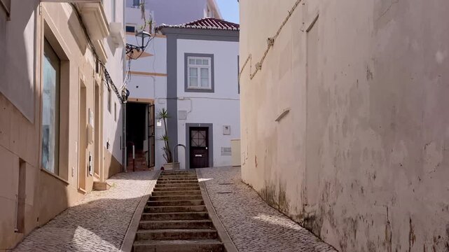 Old town street in Lagos, Portugal, featuring a narrow cobblestone path ascending with stone steps, surrounded by traditional white buildings