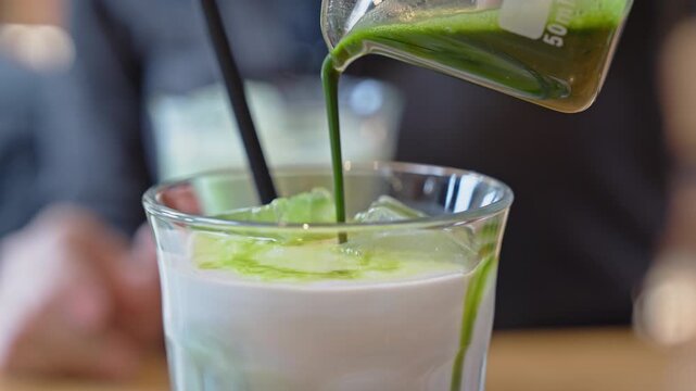 Pouring green matcha tea over ice in milk glass for latte drink cafe beverage