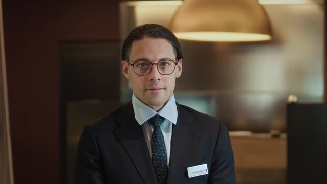 Waist up portrait of young adult Caucasian man wearing suit and glasses standing in hotel lobby looking directly at camera. Footage works well for corporate communication or hospitality marketing