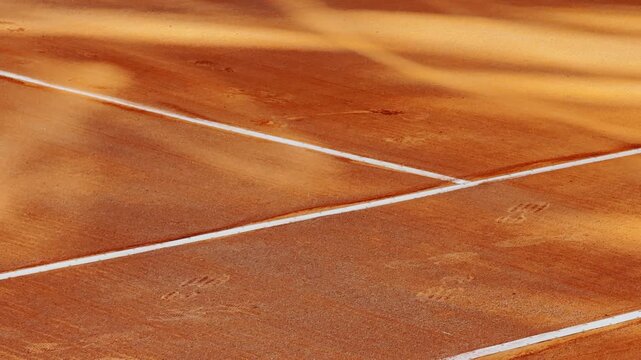Close-up of an orange clay tennis court with crisp white boundary lines, textured playing surface, rake marks, footprints, and warm sunlight.