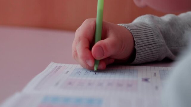 Child's hand holding green pen writes on homework sheet with colorful charts and numbers, soft lighting illuminating the workspace in a cozy indoor setting