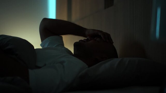 Young man lying awake in bed struggling with insomnia and depression at night. Mental health, Stress sleep disorder. Emotional exhaustion and wellness concept.
