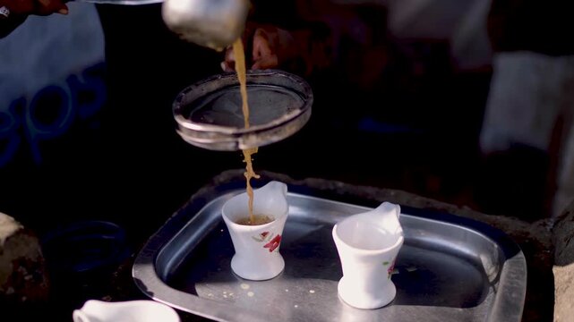 Hands pouring hot chai through metal strainer into small cups on tray at street stall in Sindh Pakistan
