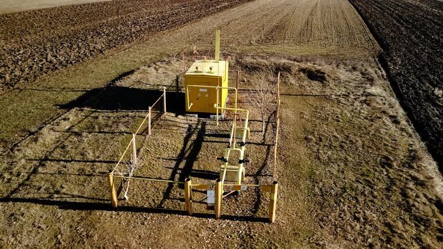 Yellow industrial gas metering station with fenced perimeter and visible piping situated in a rural landscape with plowed fields and clear sky in the background
