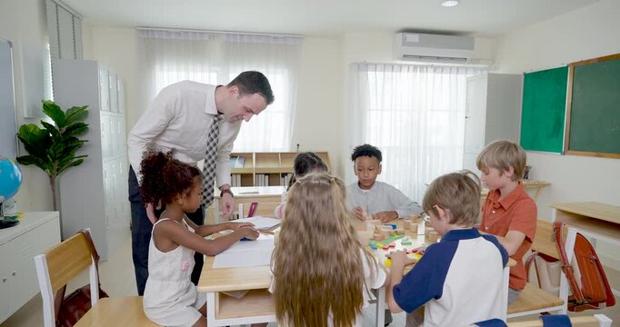 Adult male teacher moves around group table checking colorful block activity as multicultural pupils work together, classroom guidance builds cooperation, confidence, responsibility, learning routine